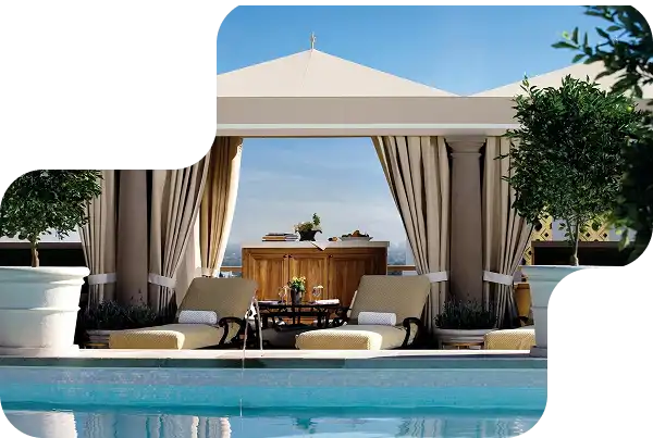 Luxury poolside cabana with beige outdoor curtains, lounge chairs, and a wooden table under a canopy.