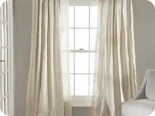 Light-colored pinch pleat curtains hanging on a large window in a bright room.