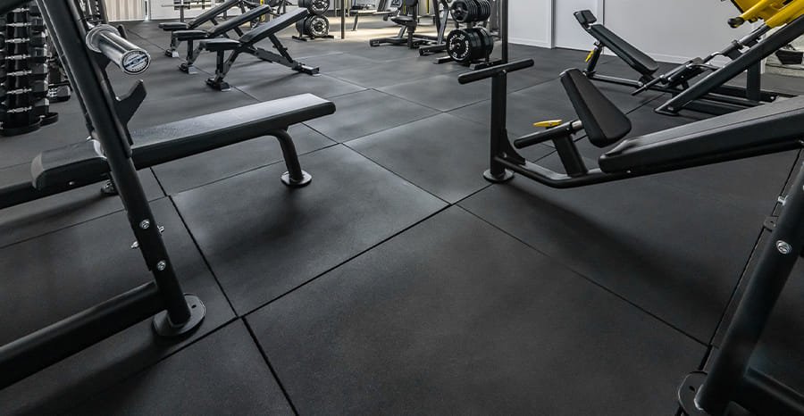 Thick black gym mats placed under benches and squat racks in a commercial gym.
