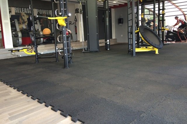 Black interlocking gym mats covering the floor of a workout area with various exercise machines.